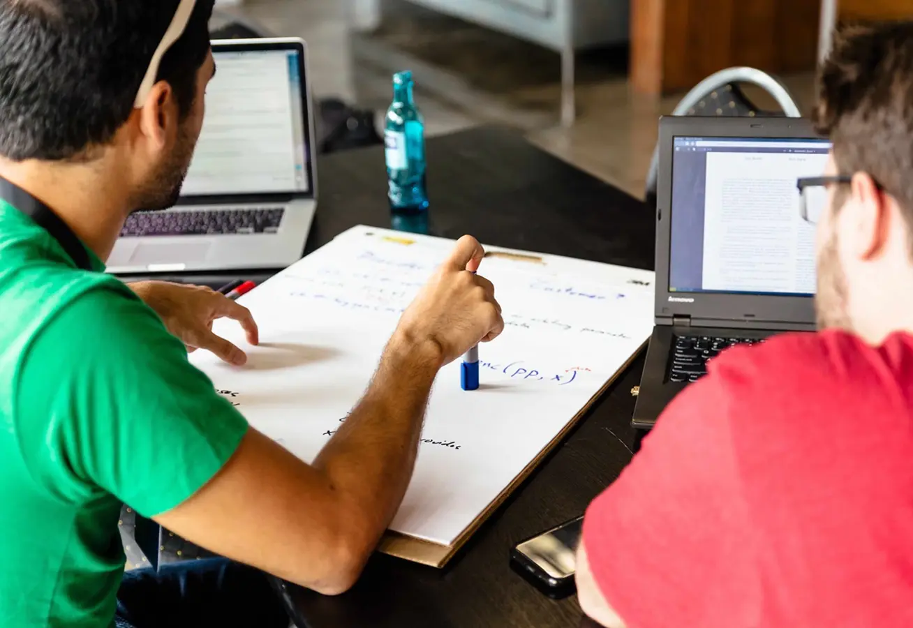 Two people sitting at a table with laptops and a large sheet of paper, collaborating and writing notes with a marker.